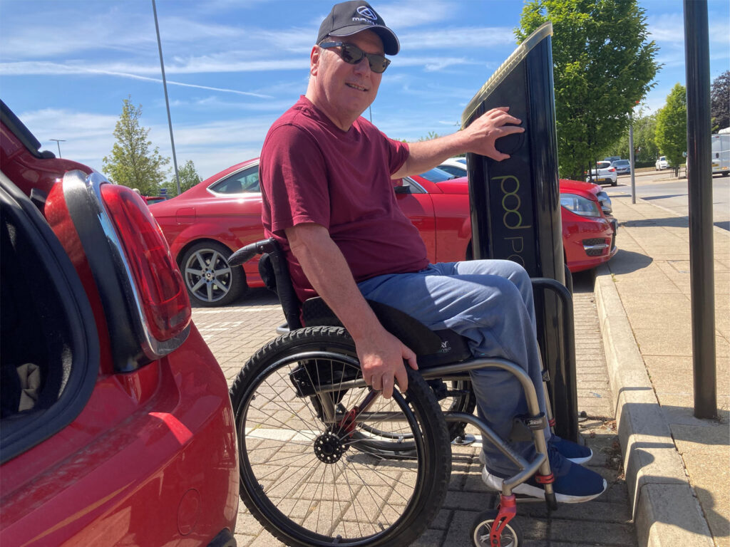 Our Accessible Vehicle and Travel Advice Advocate Roger Warner is in front of his red car in his wheelchair, with his hand on a car charging point.