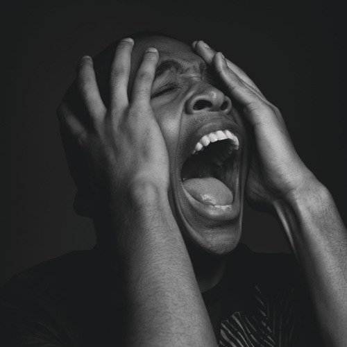 A black man has his mouth open and hands covering his head in anguish, he is in a dark room and barely lit.
