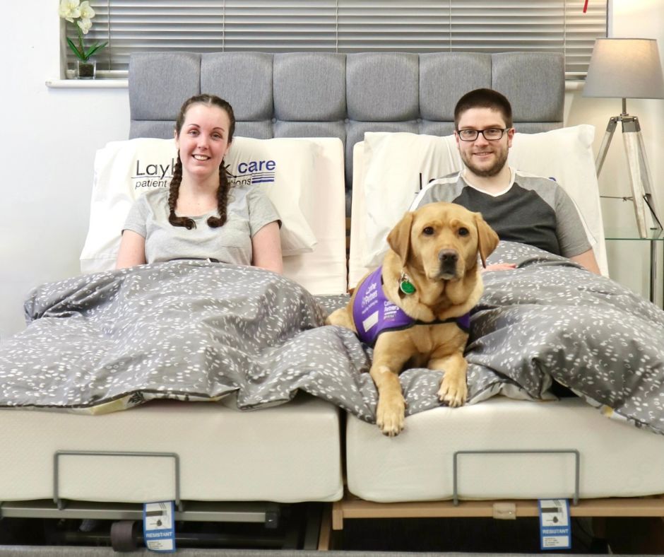 A young couple are sitting upright on the Laybrook Eyre Adjustable Bed, laying between them is their assistance dog.