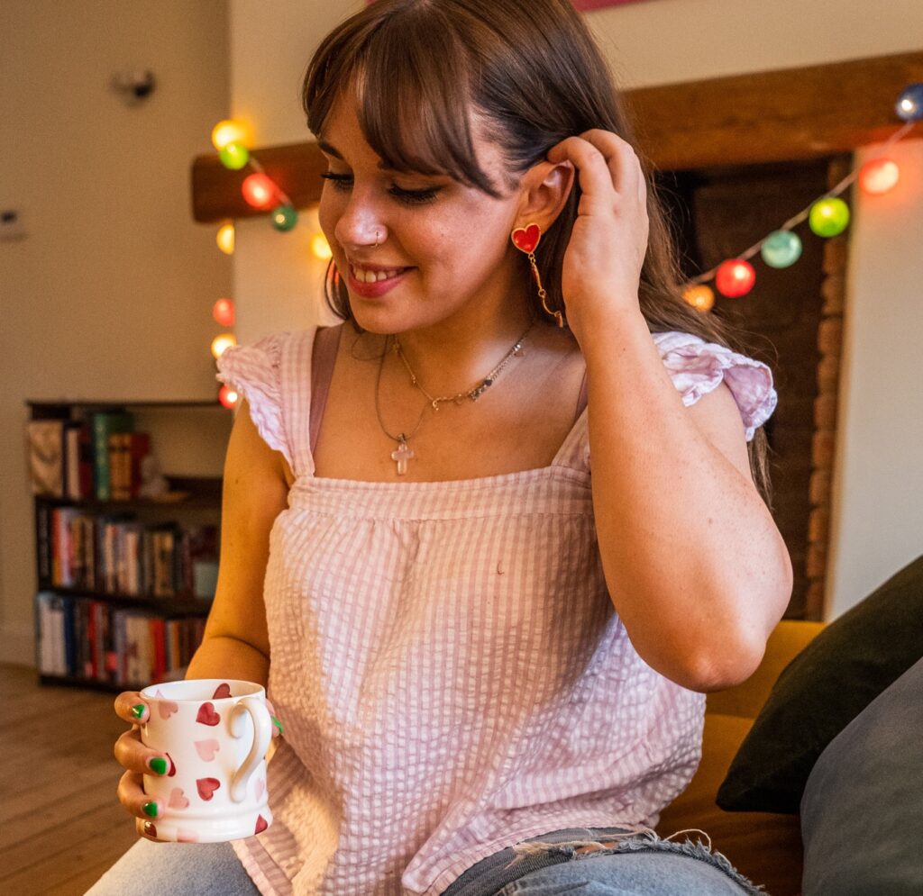 Emi Ainscough sitting in a living room with fairy lights. She is tucking her hair behind her ear and holding a mug with hearts on it.
