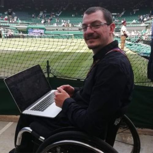 Nate Williams, People's Panel Speaker, in his wheelchair with a laptop on his lap next to a tennis court.