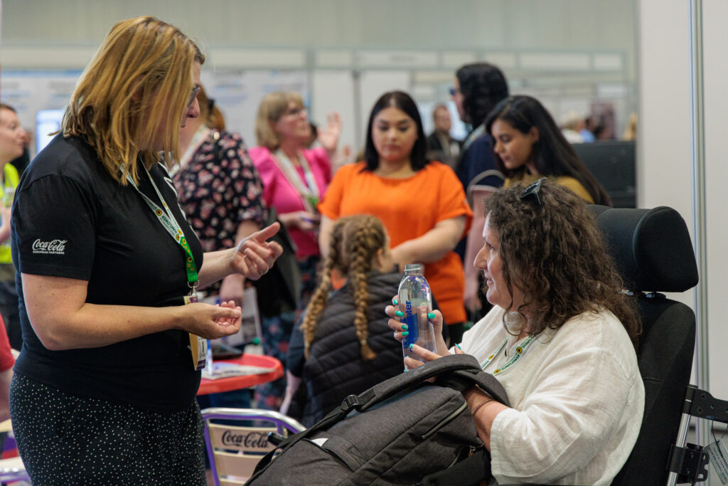 A woman in a power chair having a discussion with an exhibitor. Behind them you can see a busy exhibition hall.