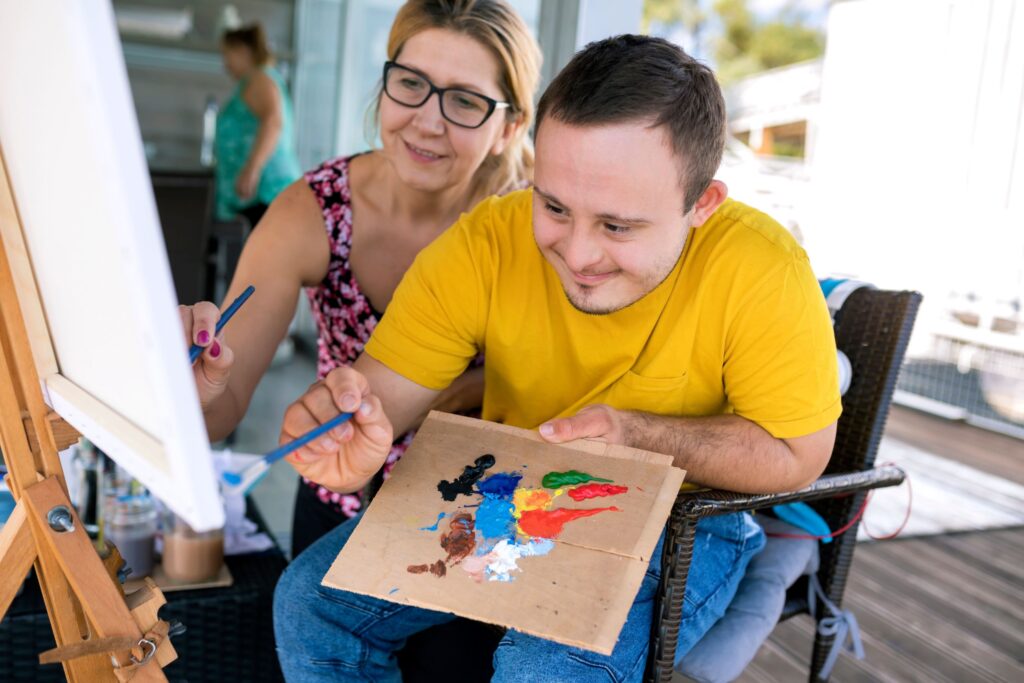 Young man and his carer are smiling and painting on a canvas. The topic of the article is disability and arts.