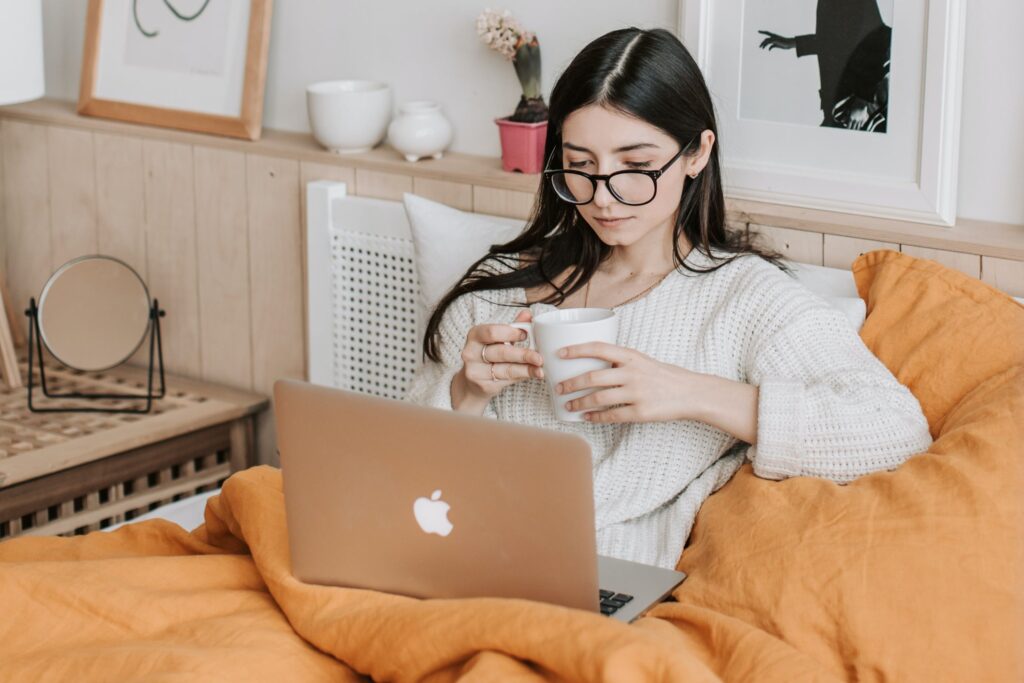 A young woman is lying propped up in bed with a cup of tea and looking at her laptop screen. The article is about digital accessibility for people with a disability.