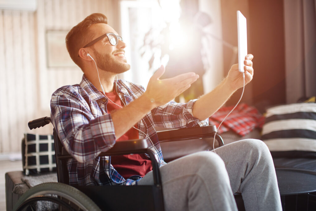 Assistive technology in home living: young man in wheelchair using tablet while being at home. He is gesturing during video call.