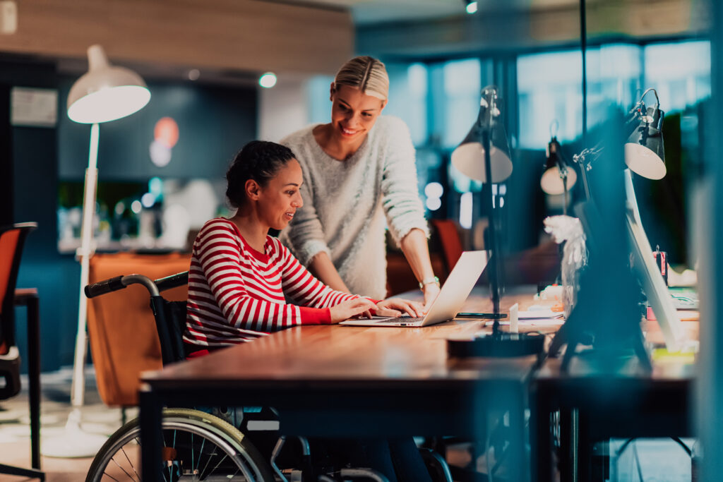 Businessperson in a wheelchair working in a creative office. Business team in modern coworking office space. Colleagues working in the background at late night. Image illustrating workplace mental health article.